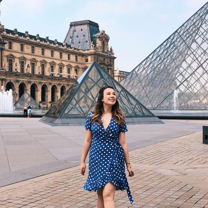Pregnant Disney superfan smiling in a blue polka dot dress near glass pyramid structures at a historic museum courtyard. Pregnant Disney superfan smiling in a blue polka dot dress near glass pyramid structures at a historic museum courtyard.