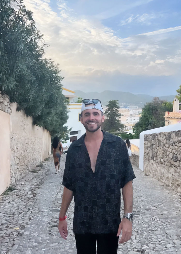 Young man smiling outdoors during hair transplant recovery, wearing a patterned shirt and white cap on a cobblestone street. Young man smiling outdoors during hair transplant recovery, wearing a patterned shirt and white cap on a cobblestone street.