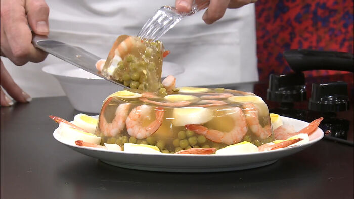 A slice of shrimp jello being served from a gelatin mold with peas, shrimp, and boiled eggs on a plate. A slice of shrimp jello being served from a gelatin mold with peas, shrimp, and boiled eggs on a plate.