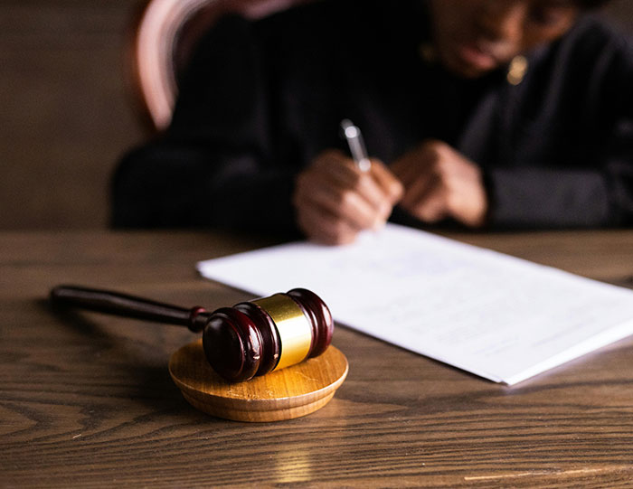 Gavel lying on wooden table in courtroom setting with blurred person writing legal document in background. Gavel lying on wooden table in courtroom setting with blurred person writing legal document in background.