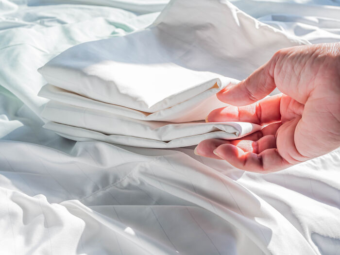 Hand folding a white bedsheet on a bed, illustrating a so-called genius life hack that adds extra work.