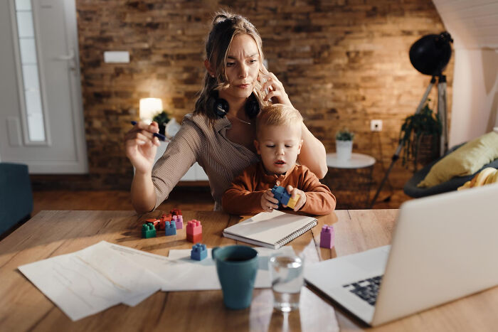 Mother multitasking with child, phone, and work at home, illustrating unfair relationships due to gender roles in parenting.