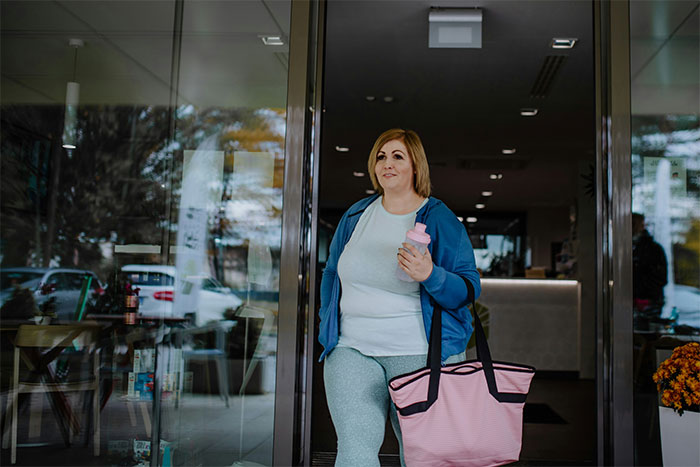 Overweight woman at gym entrance holding water bottle and bag, reflecting on childhood trauma and past bullying experiences. Overweight woman at gym entrance holding water bottle and bag, reflecting on childhood trauma and past bullying experiences.
