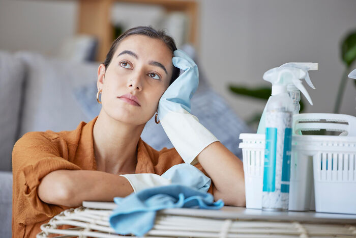 Young woman in rubber gloves resting tiredly on a table with cleaning supplies, illustrating unfair gender roles in relationships.