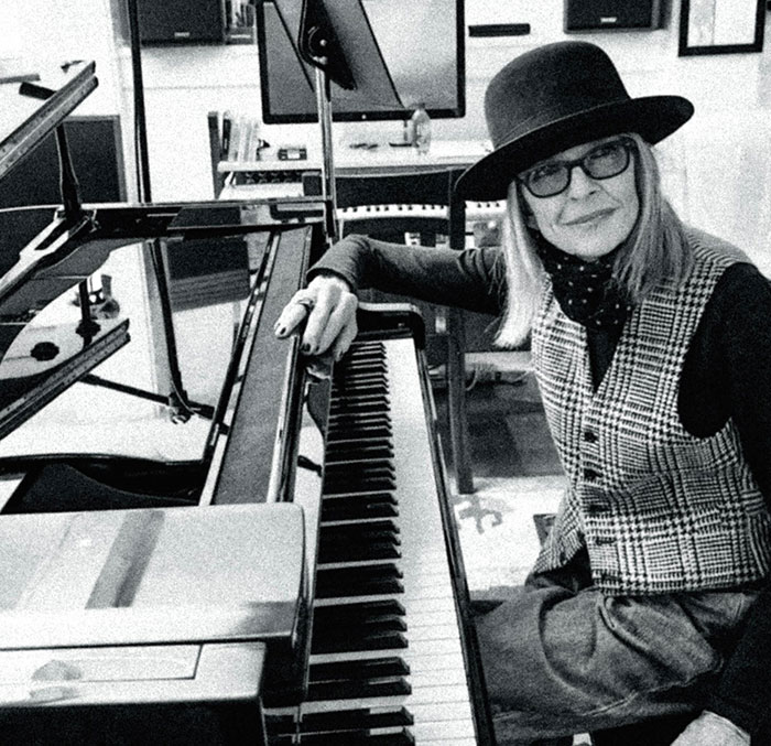 Black and white photo of Diane Keaton sitting at a piano, wearing a hat and glasses in a cozy indoor setting. Black and white photo of Diane Keaton sitting at a piano, wearing a hat and glasses in a cozy indoor setting.
