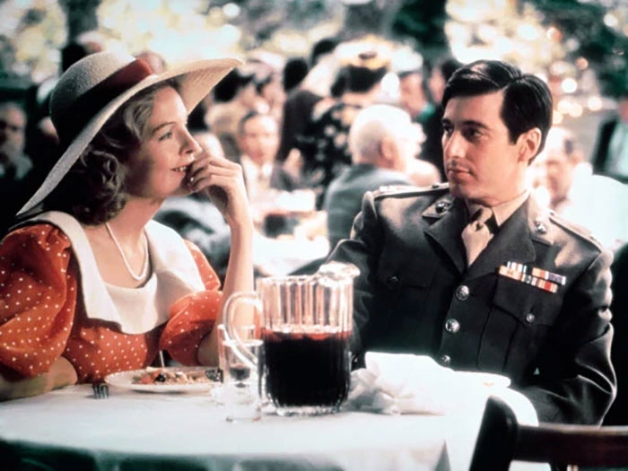 Scene with Diane Keaton and a man in military uniform seated at an outdoor table, vintage setting reflecting a past era. Scene with Diane Keaton and a man in military uniform seated at an outdoor table, vintage setting reflecting a past era.