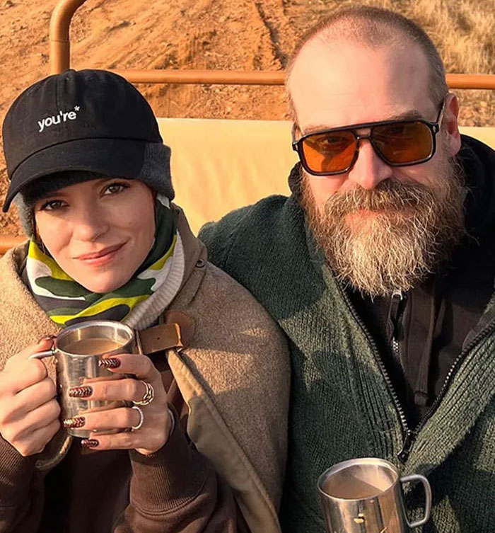 David Harbour and a younger woman sitting outdoors, holding metal mugs during a casual moment. David Harbour and a younger woman sitting outdoors, holding metal mugs during a casual moment.