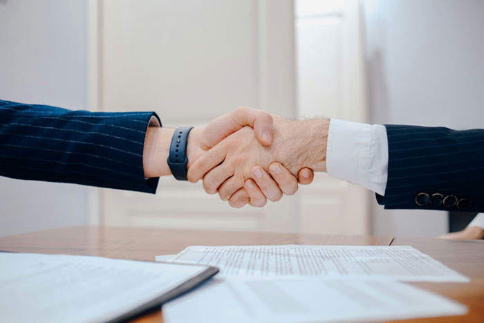 Two professionals shaking hands over a desk with documents, illustrating company claims about raises and new hire salary. Two professionals shaking hands over a desk with documents, illustrating company claims about raises and new hire salary.