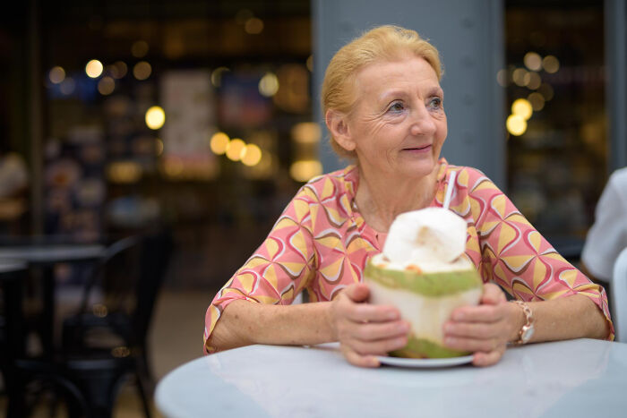 Elderly woman at a restaurant, holding a coconut drink, illustrating a waiter’s painful table for two moment ending as table for one.