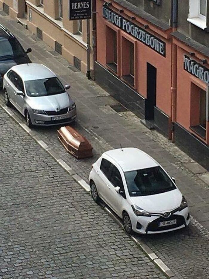 Confusing image of a coffin placed on a sidewalk between parked cars, creating a strange and weird street scene.