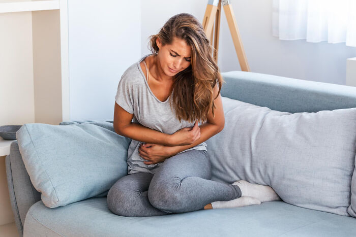 Young woman sitting on a couch, holding her stomach in pain, illustrating terrifying medical conditions without symptoms.