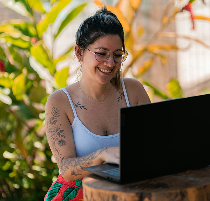 Woman with tattoos smiling and using a laptop outdoors, reflecting laughter and guilt after hot revenge on neighborhood kids.