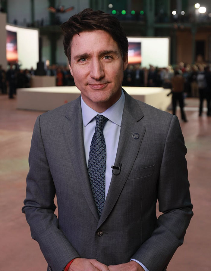 Justin Trudeau in a grey suit speaking at an indoor event with people and screens in the background. Justin Trudeau in a grey suit speaking at an indoor event with people and screens in the background.