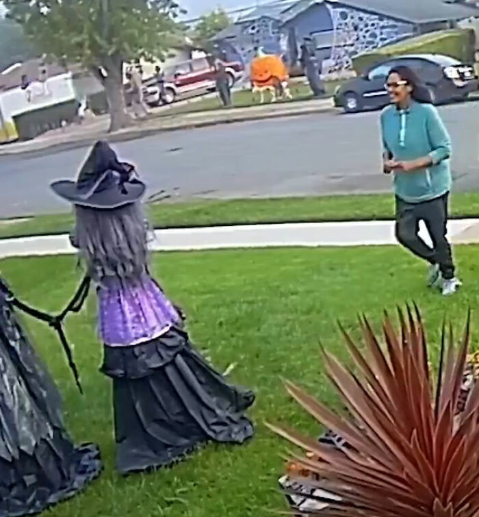 Woman in blue hoodie near Halloween witches lawn decorations on a suburban street during daytime.