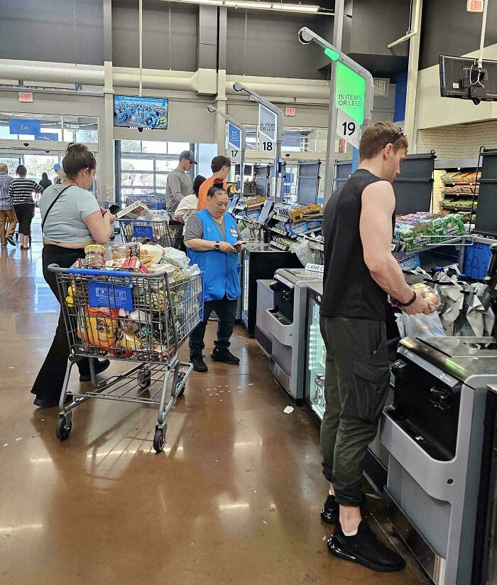 Customers and employees at self-checkout lanes inside a Walmart store with full shopping carts and bags.