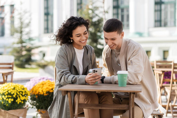 Two young people sitting at an outdoor table smiling and looking at a phone, surrounded by flowers and buildings, creepy things context.