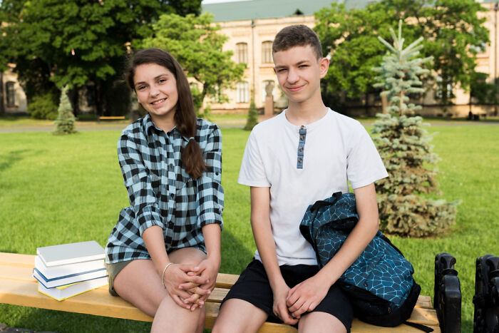 Two teenagers sitting on a bench outdoors with backpacks and books, representing popular kids in school scenarios.