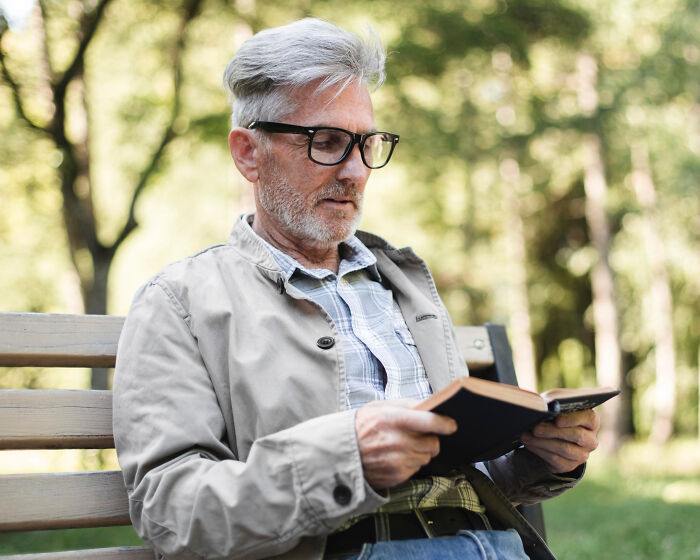 Elderly man wearing glasses sitting on a park bench reading a book, representing a patient with dementia.