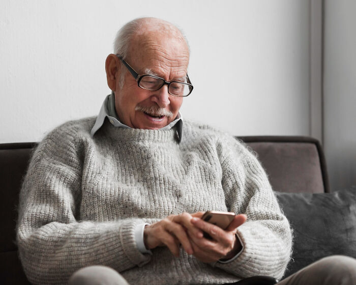 Elderly man with glasses smiling while struggling with technology skills on his smartphone at home.