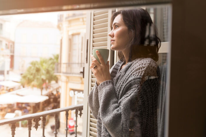 Woman in a gray sweater holding a cup, looking thoughtfully outside a window reflecting disturbing and creepy thoughts.