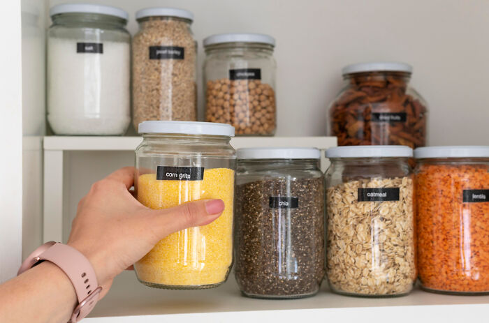 Hand reaching for labeled glass jar of corn grits on pantry shelf with other jars in organized kitchen storage life hacks.