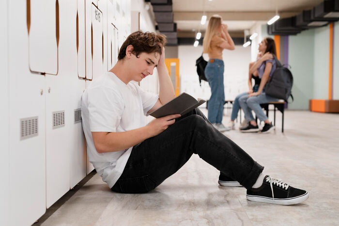 Teen boy sitting alone by lockers reading a book while popular kids talk in the background in school hallway outcasts