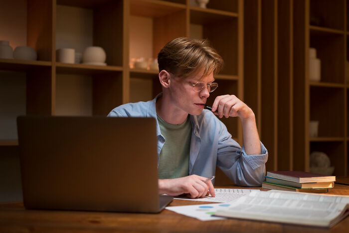 Young man wearing glasses working late at home, thinking deeply about gender roles in unfair relationships.
