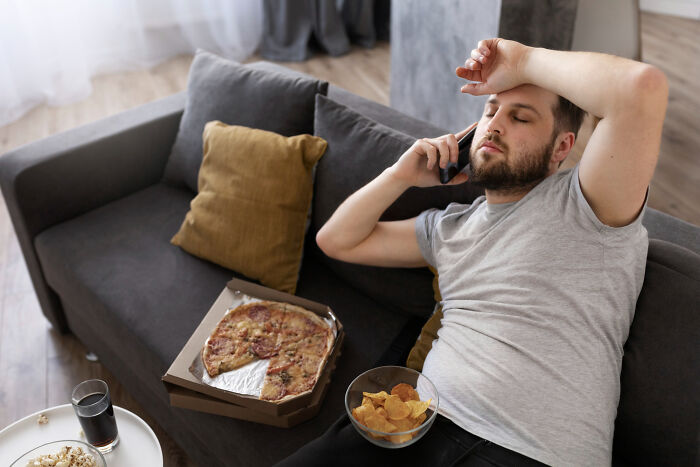 Man on couch with pizza and chips, appearing exhausted, illustrating unfair relationships due to gender roles.