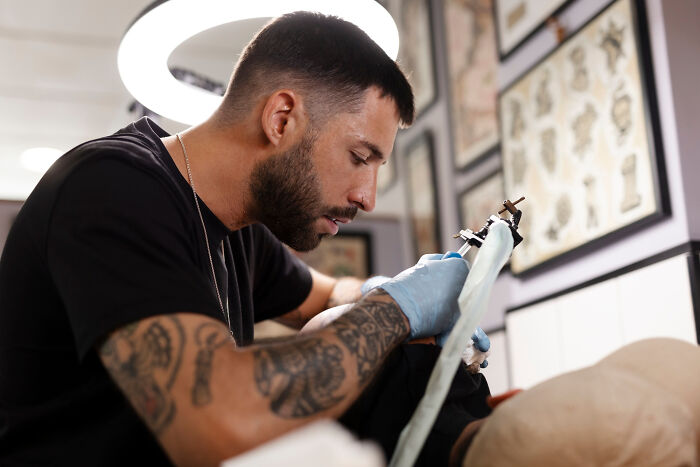 Tattoo artist with sleeve tattoos focused on drawing a design with a machine under ring light in tattoo studio.