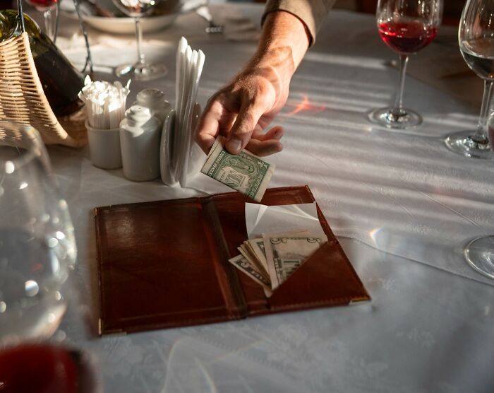 Hand placing cash in a leather bill holder on a restaurant table with wine glasses around the scene.