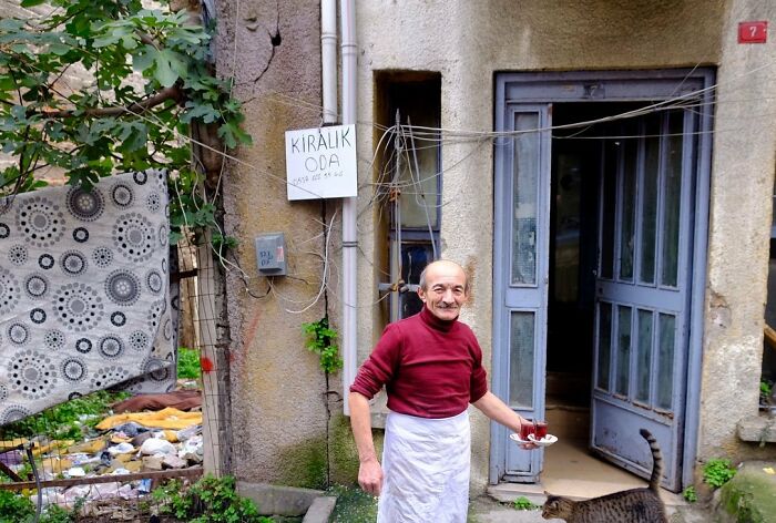 Elderly man holding tea glass outside old Istanbul street building with a cat walking nearby on the worn pathway.