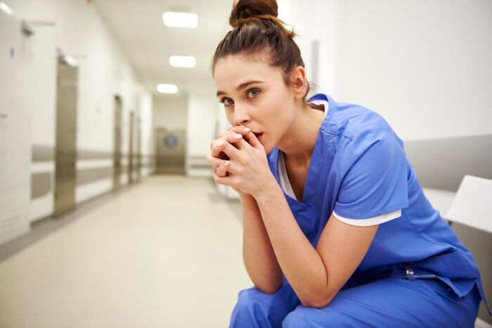 Nurse in blue scrubs sitting in hospital hallway looking worried, evoking scary ghost stories hospital work atmosphere.