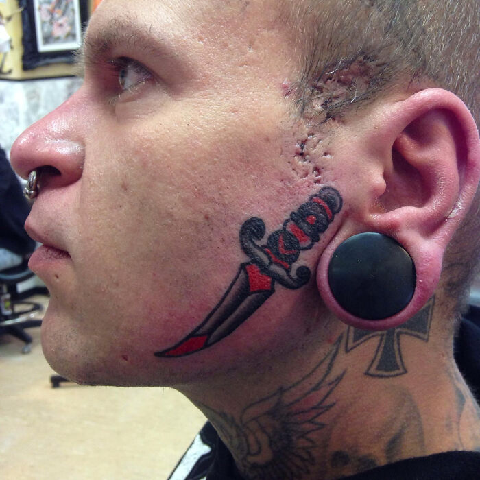 Close-up of a man's face showing a bold red and black face tattoo with facial piercings and stretched earlobe plug.