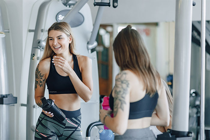 Two young women with tattoos in athletic wear smiling and talking at the gym during a workout session. Two young women with tattoos in athletic wear smiling and talking at the gym during a workout session.