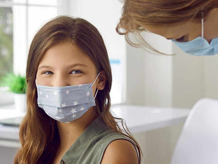 12-year-old girl wearing a star-patterned mask sitting in a medical office with a masked adult attending to her. 12-year-old girl wearing a star-patterned mask sitting in a medical office with a masked adult attending to her.