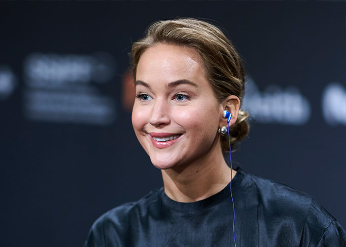 Jennifer Lawrence smiling during a public event wearing a black top and blue in-ear headphones discussing plastic surgery plans. Jennifer Lawrence smiling during a public event wearing a black top and blue in-ear headphones discussing plastic surgery plans.