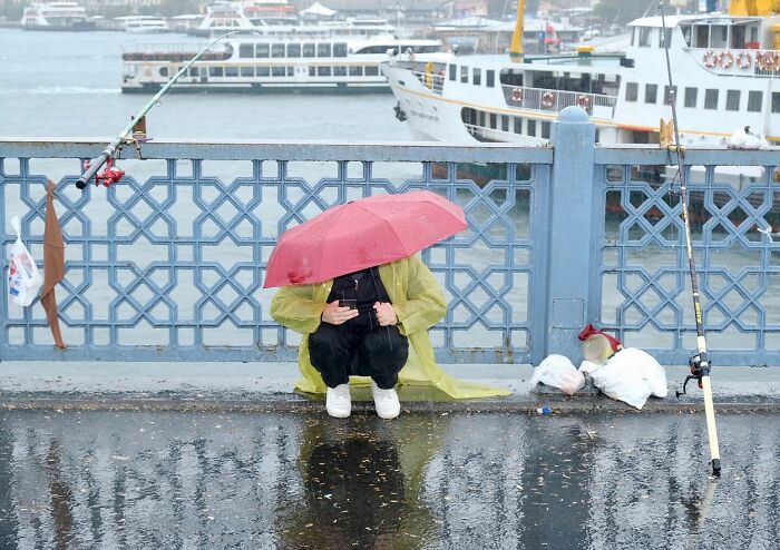 Person fishing on Istanbul streets under a red umbrella on a rainy day with boats and wet pavement visible nearby.