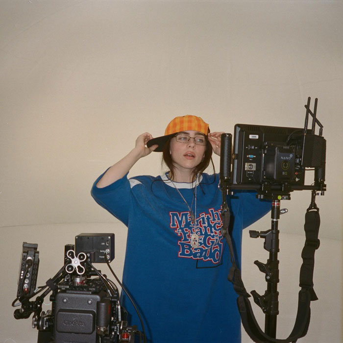 Billie Eilish on set wearing an orange cap and blue sweatshirt amid professional filming equipment and cameras. Billie Eilish on set wearing an orange cap and blue sweatshirt amid professional filming equipment and cameras.