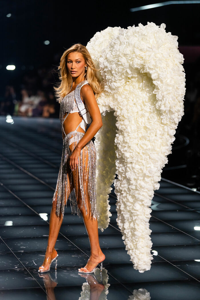 Bella Hadid walking runway wearing large white Victoria’s Secret angel wings with silver fringe outfit on glossy catwalk. Bella Hadid walking runway wearing large white Victoria’s Secret angel wings with silver fringe outfit on glossy catwalk.
