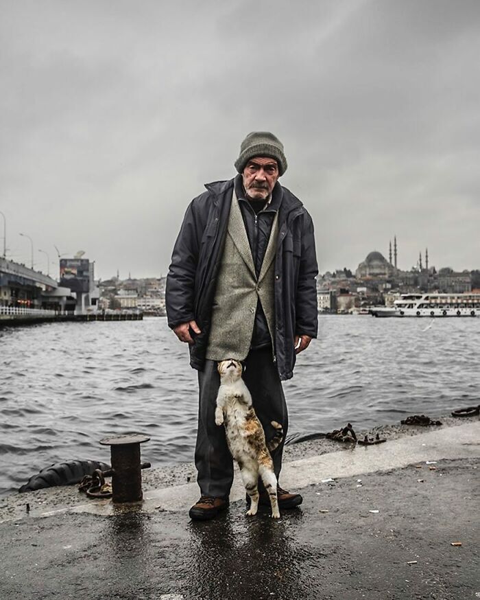 Elderly man standing by the water in Istanbul streets with a cat rubbing against his legs on a cloudy day.