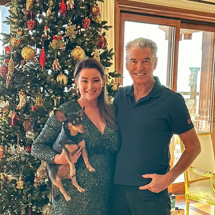 Pierce Brosnan’s wife in a sparkling dress holding a small dog, standing beside him by a decorated Christmas tree indoors. Pierce Brosnan’s wife in a sparkling dress holding a small dog, standing beside him by a decorated Christmas tree indoors.