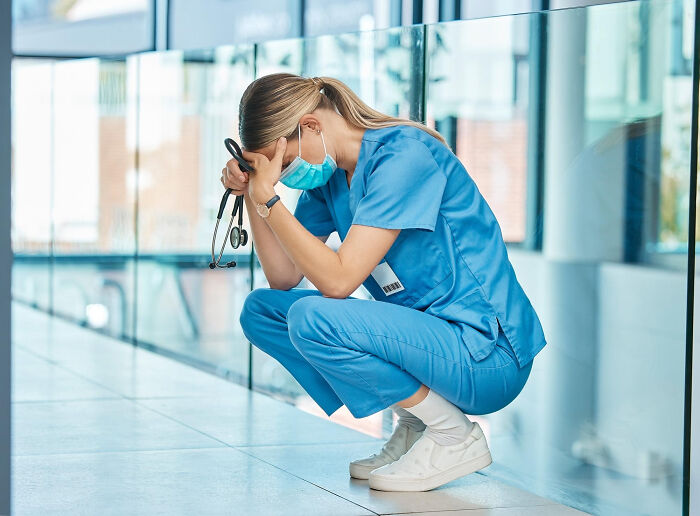 Nurse in blue scrubs crouching with head in hands, wearing mask, capturing the mood of scary ghost stories in hospital work.