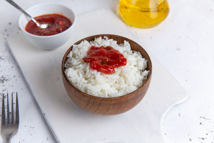Bowl of white rice topped with ketchup, next to a small bowl of red sauce and a bottle of oil on a white surface.