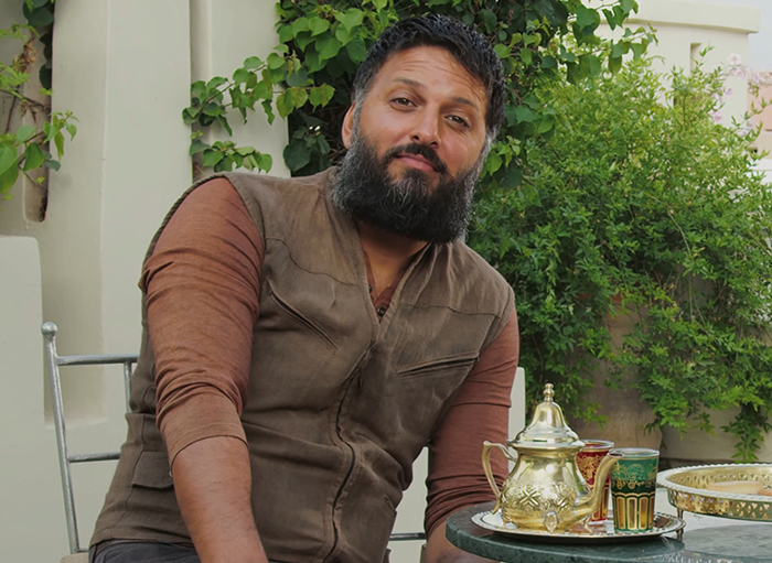 Man with a beard and brown vest sitting outdoors at a table with a golden teapot and colorful cups, greenery behind. Man with a beard and brown vest sitting outdoors at a table with a golden teapot and colorful cups, greenery behind.
