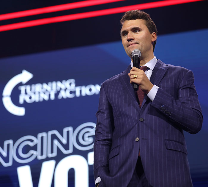 Charlie Kirk speaking at Turning Point Action event, wearing a striped suit and holding a microphone on stage. Charlie Kirk speaking at Turning Point Action event, wearing a striped suit and holding a microphone on stage.