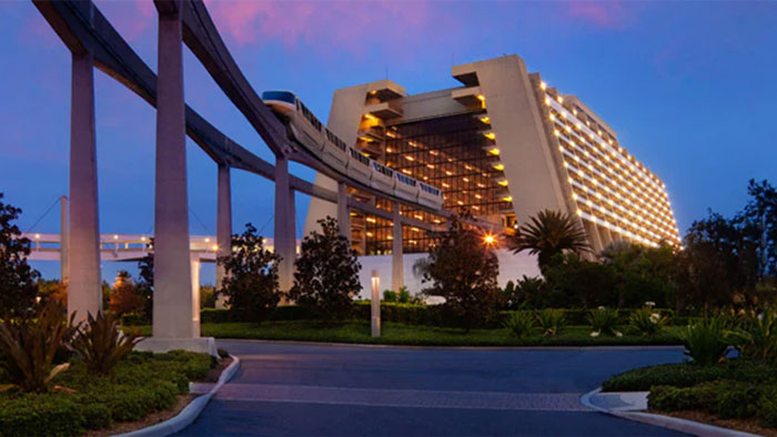 Monorail passing in front of a brightly lit Disney resort building at dusk, surrounded by trees and landscaping. Monorail passing in front of a brightly lit Disney resort building at dusk, surrounded by trees and landscaping.