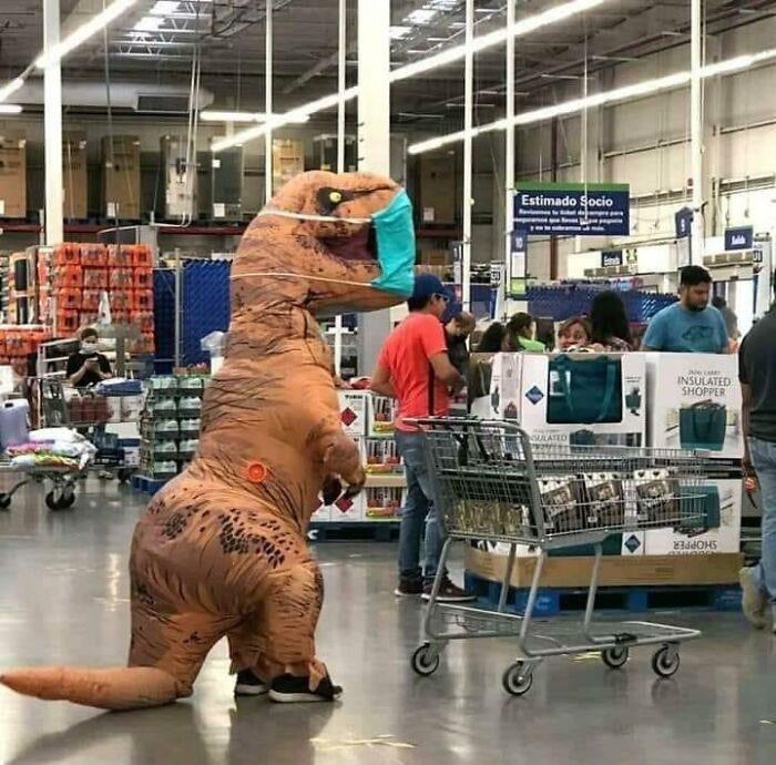 Person in inflatable dinosaur costume wearing a mask while shopping inside a busy Walmart store with carts and other shoppers.