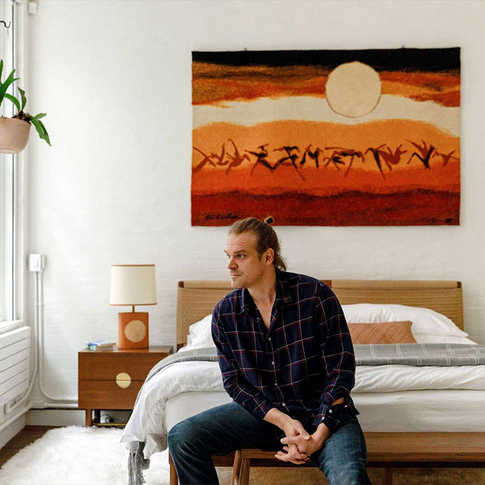 Man sitting on a bed in a modern bedroom, with a large colorful tapestry on the wall, reflecting on Lily Allen's album. Man sitting on a bed in a modern bedroom, with a large colorful tapestry on the wall, reflecting on Lily Allen's album.
