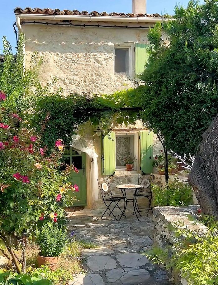 Charming stone cottage with green shutters surrounded by lush gardens and vibrant flowering plants in a peaceful setting.