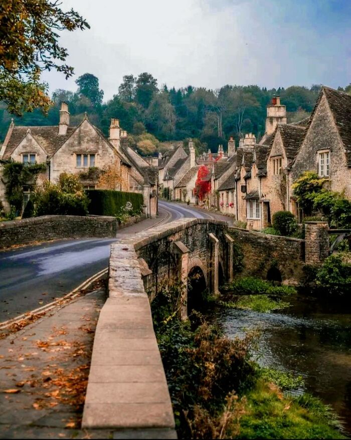 Stone cottages and gardens along a winding road with a historic stone bridge over a river in a peaceful village setting.